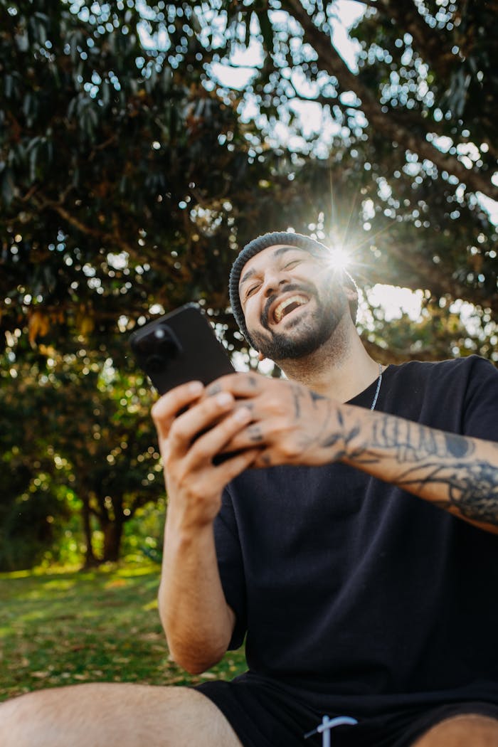 A cheerful man with tattoos laughing while holding a smartphone outdoors in a sunny park.