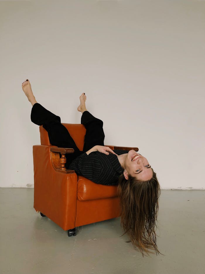 A woman joyfully reclines upside down in a vintage leather armchair, showcasing a carefree moment indoors.